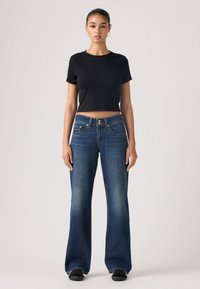 Νεαρή γυναίκα που στέκεται όρθια, φορώντας ένα μαύρο κοντό t-shirt, μπλε παντελόνι καμπάνα και μαύρα παπούτσια μπροστά σε απλό φόντο.