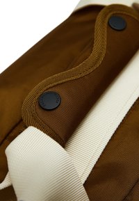 Bolsa de nylon marrón con acentos en color crema, que presenta una superficie texturizada, costuras en contraste y un detalle de hardware negro en forma circular.