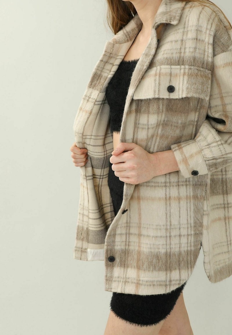 Beige plaid shirt jacket made of soft fabric, featuring black buttons and a relaxed fit. Inner lining visible with contrasting texture.