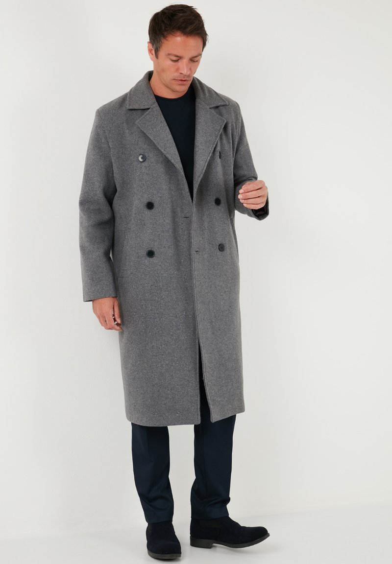 Grauer Wollmantel mit zweireihigem Design, großen schwarzen Knöpfen und Spitzrevers. Hat einen geraden Schnitt und reicht bis zur Mitte der Wade.