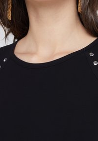 Top noir avec un col rond, texture lisse et accents métalliques aux épaules. Design simple et minimaliste.