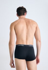Mann, der mit dem Rücken zum Betrachter steht, trägt schwarze Calvin Klein Boxer Shorts vor einem schlichten weißen Hintergrund.