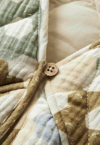 Puitnupp lähivõte, kinnitatud kapuutsitud kangale, millel on rohelised, pruunid ja beežid maskeerimismustrid ning nähtavad õmblused.