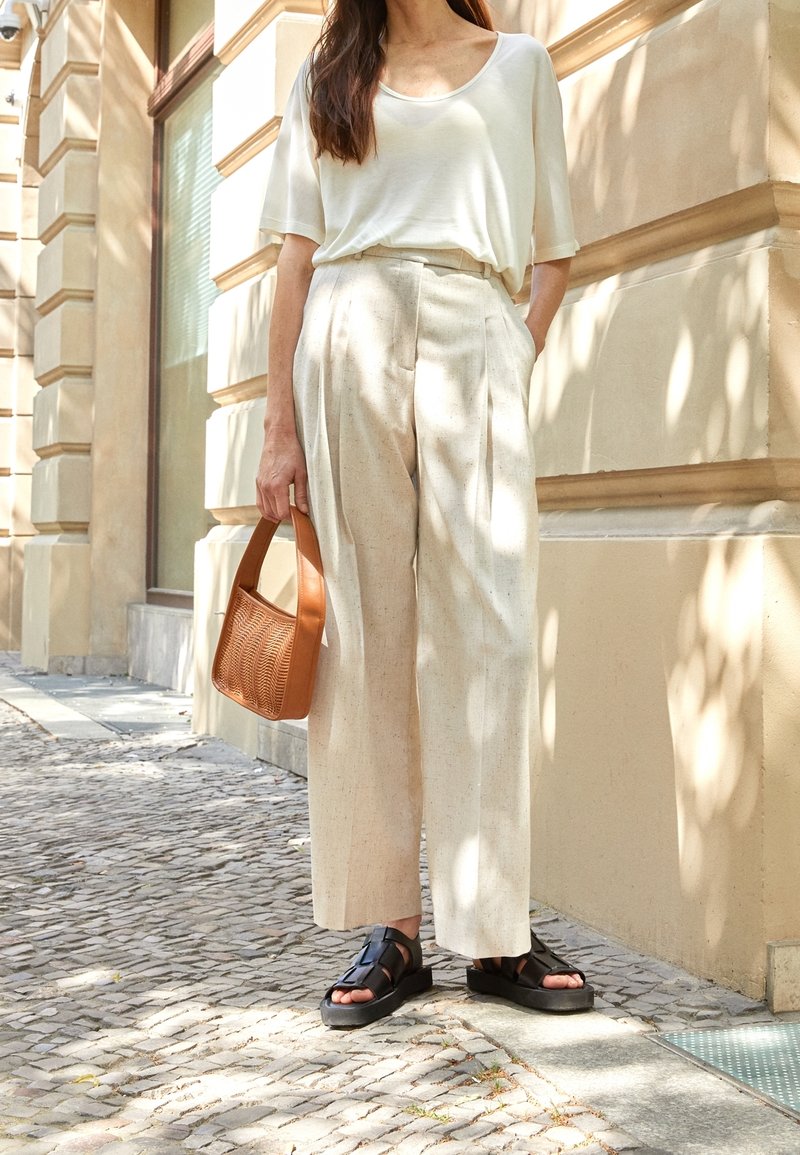 Lehká krémová košile, béžové plisované kalhoty, černé sandály a texturovaná hnědá taška. Stojí u světlé zdi a na kostkované dlažbě.