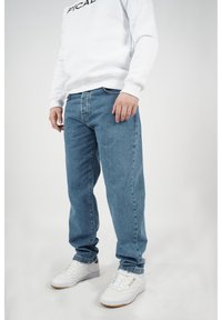 Man i vit sweatshirt, blå jeans och vita Reebok-sneakers, stående mot en ljusgrå bakgrund.