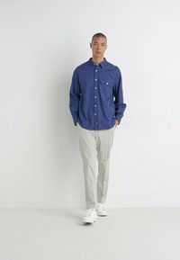 Camisa de botones de algodón azul oscuro, con un bolsillo en el pecho; combinada con pantalones sueltos de color claro y zapatillas blancas.