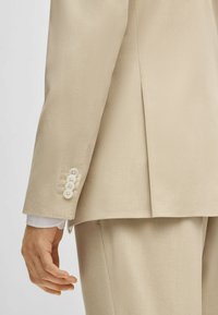 Manica di giacca beige con quattro bottoni bianchi, texture liscia e design su misura. Cucitura visibile lungo il braccio e la spalla.
