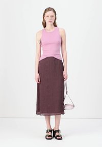 Rosa gerippter Tanktop, kombiniert mit einem knielangen, braunen, plissierten Rock. Das Model steht in schwarzen Sandalen mit Schnallen und hält eine kleine hellrosa Tasche.