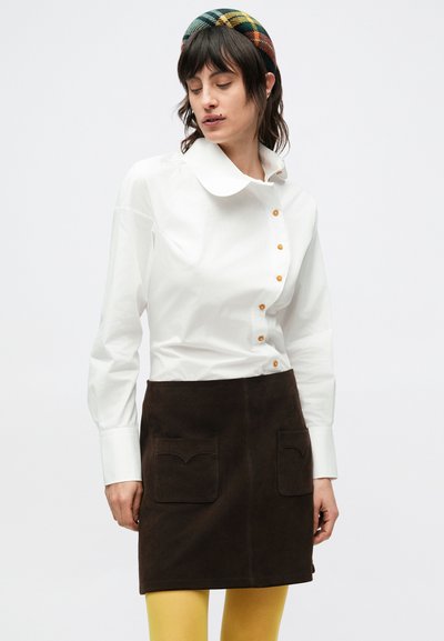 Biela košeľa na gombíky s veľkým golierom a oranžovými gombíkmi, doplnená hnedou semišovou sukňou s dvoma vreckami vpredu. Jasne žlté pančuchy.