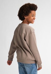 Felpa marrone con collo rotondo, maniche lunghe, polsini e bordo a coste, indossata da un bambino rivolto leggermente di spalle, abbinata a jeans blu.