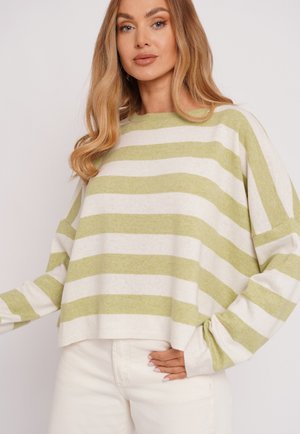 Kobieta o długich blond włosach, ubrana w luźny sweter w zielono-białe paski oraz białe spodnie na jednolitym tle.