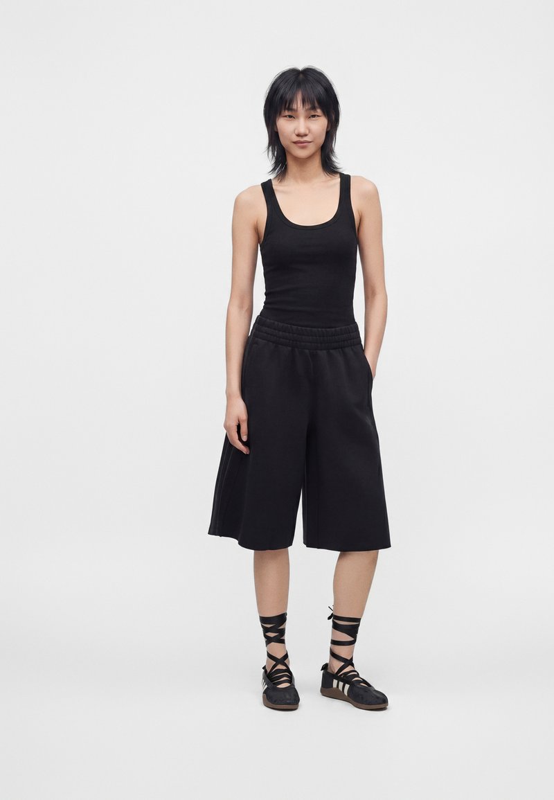 Femme debout portant un top noir sans manches, un short large noir longueur genoux et des chaussures noires à lacets avec des brides sur fond blanc.