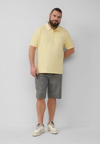 Gelbes Poloshirt mit kurzen Ärmeln, graue Shorts und weiße Sneakers mit dunklen Akzenten. Der Stoff wirkt glatt und leicht.