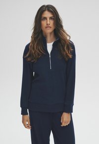 Femme avec de longs cheveux bruns portant un sweat-shirt à demi-zip côtelé bleu marine et un pantalon assorti, se tenant contre un fond clair.