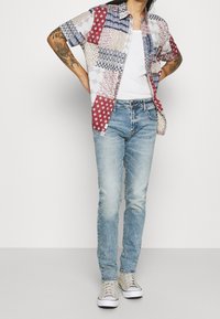 Chemise à manches courtes avec motifs multiples en tons rouge, bleu et blanc ; associée à un jean bleu clair et des baskets grises.