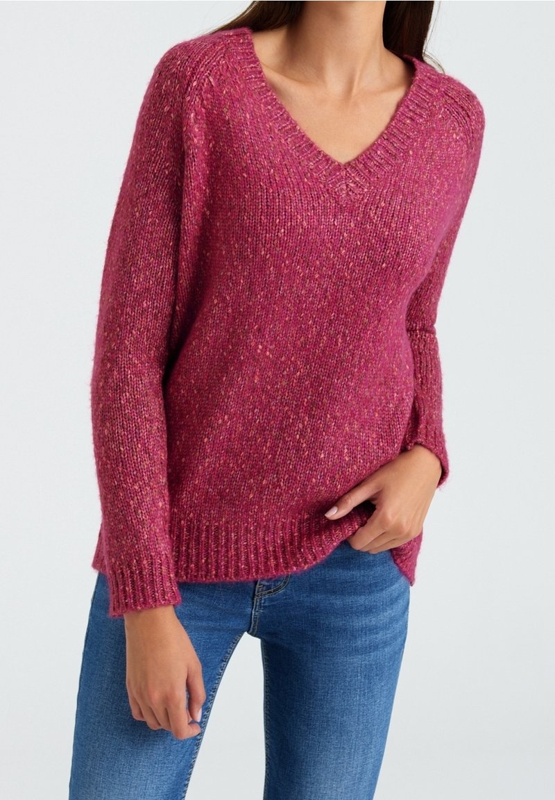 Femme portant un pull texturé à col en V rose framboise et un jean bleu, avec une main reposant dans sa poche, sur un fond uni.