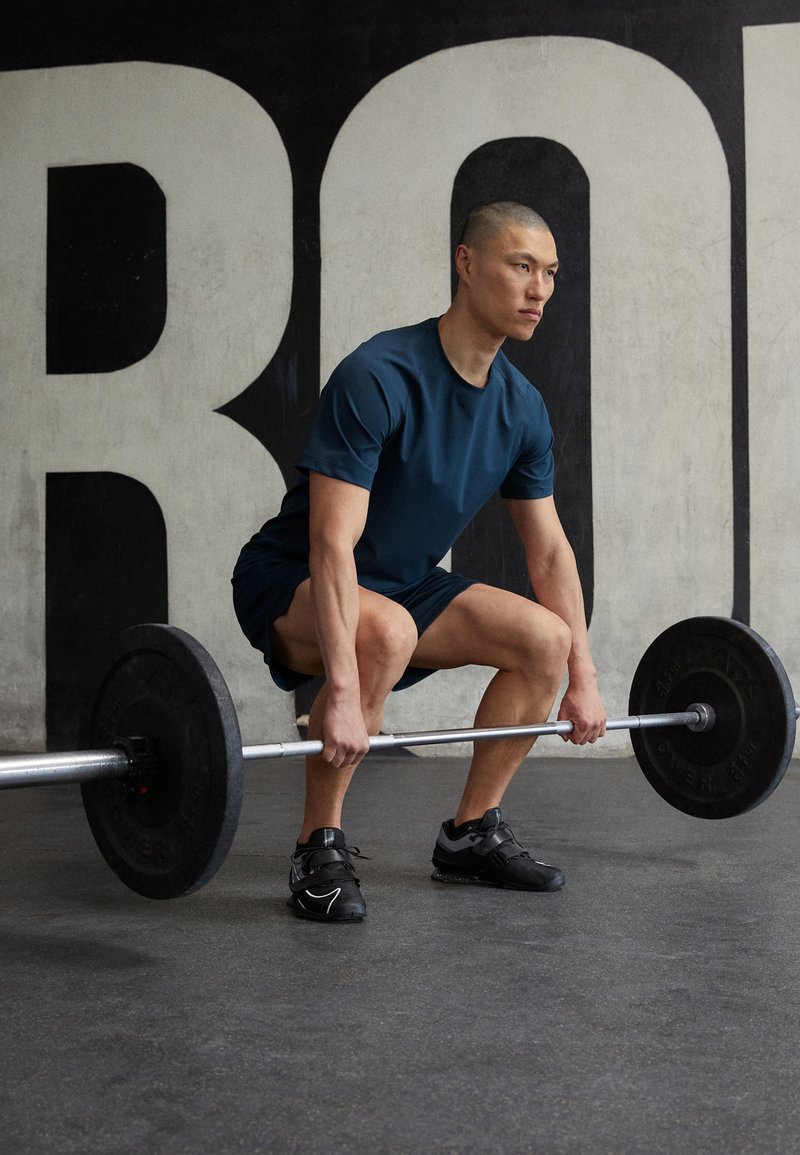Osoba w granatowej koszulce z krótkim rękawem i czarnych szortach podnosi sztangę z czarnymi ciężarami w siłowni, opierając się o betonową ścianę.