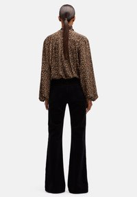 Donna con coda di cavallo lunga, indossa una camicetta con stampa leopardata e pantaloni neri a gamba larga a vita alta, in piedi di spalle su uno sfondo bianco.