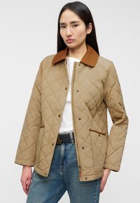 Μπεζ μπουφάν με καπιτονέ σχέδιο και καφέ γιακά, με κουμπώματα πατς και δύο μπροστινές τσέπες, φορεμένο πάνω από λευκή μπλούζα και μπλε τζιν.