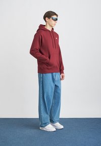 Burgundský mikina s kapucňou a klokánkovým vreckom, skombinovaná so svetlomodrými oversized džínsami a bielymi teniskami; model má na sebe modré slnečné okuliare.