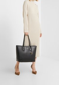 Sac cabas en cuir noir avec deux poignées, orné d'un pendentif doré et de détails métalliques. Texture lisse avec un design structuré.