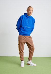 Blå huva jacka med halv dragkedja och känguruficka. Bruna sweatpants med gula bokstäver. Vita sneakers med bruna accenter. Grön golv.