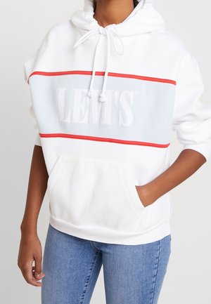 Personne portant un sweat à capuche blanc Levi's avec un panneau bleu clair et des rayures rouges, associé à un jean bleu, main dans la poche de la capuche.