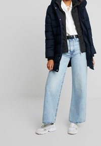 Mörkblå puffjacka med oversize-passform, långa ärmar, framdragkedja, i kombination med ljusblå vida jeans och vita sneakers.