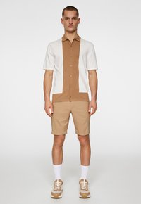 Camicetta a maniche corte con blocco di colore marrone e bianco, chiusura frontale con bottoni; abbinata a pantaloni corti beige e sneakers color pelle. Calzini bianchi visibili.