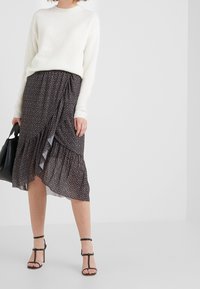 Witte gebreide trui gecombineerd met een donkere rok met bloemenpatroon en een gerimpelde zoom, en zwarte sandalen met hakken en bandjes. Zwarte tote bag in de hand.