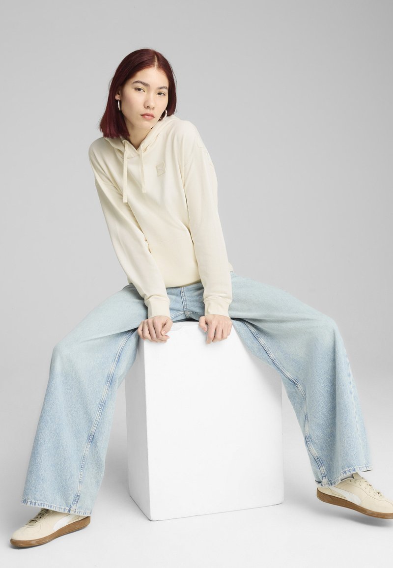 Sudadera crema clara con cordón, que presenta un pequeño logo. Combinada con jeans de pierna ancha en azul claro y zapatillas crema, sentada sobre un cubo blanco.