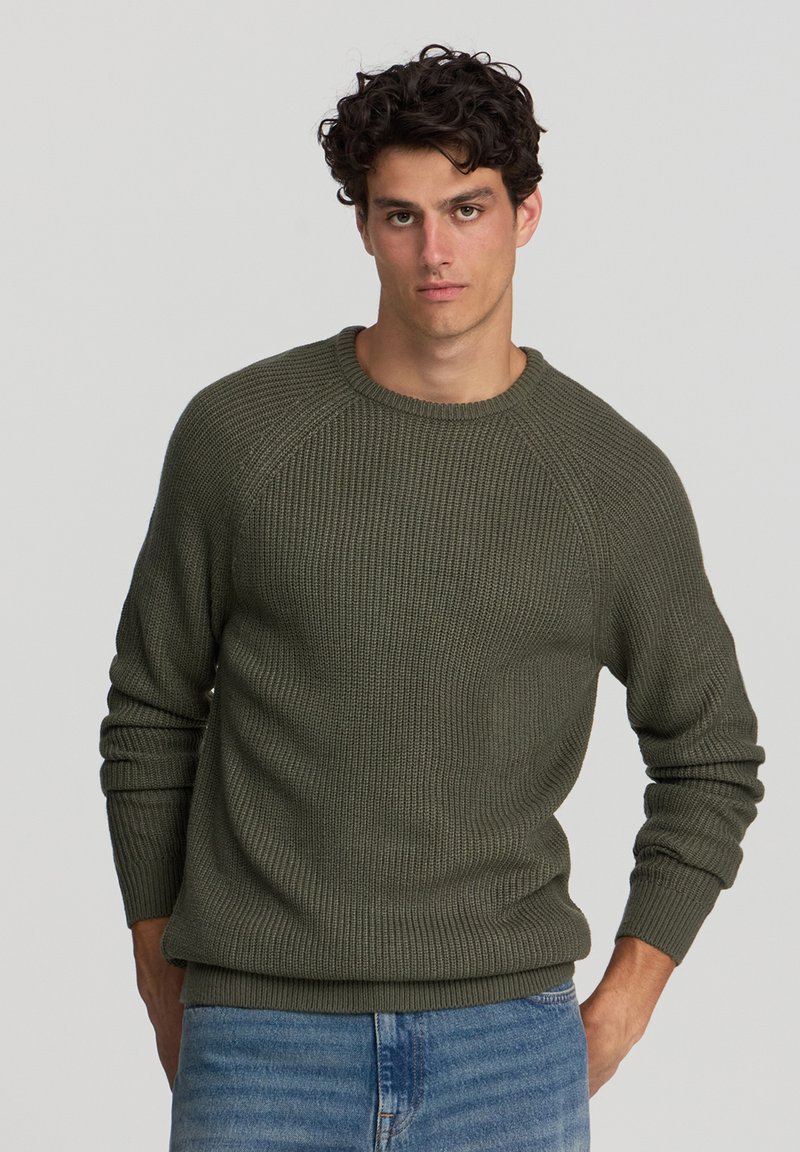 Maglione lavorato a maglia verde oliva con scollatura a girocollo, texture a coste e maniche lunghe, abbinato a jeans di denim azzurro chiaro.