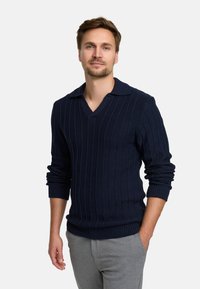 Dunkelblauer Strickpullover mit gerippter Textur, V-Ausschnitt und kurzem Kragen; enganliegende Ärmel mit umgeschlagenen Bündchen; graue Hose.