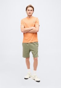 Νεαρός άνδρας στέκεται με σταυρωμένα τα χέρια, φορώντας πορτοκαλί κοντομάνικο πουκάμισο, χακί σορτς, λευκές κάλτσες και λευκά αθλητικά παπούτσια σε ανοιχτό φόντο.