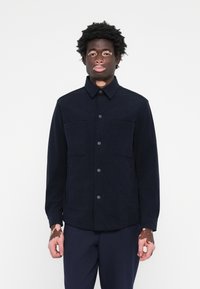 Chaqueta de trabajo azul marino en tejido estructurado. Cuenta con un cierre de botón, dos bolsillos en el pecho y un cuello. Textura suave, diseño minimalista.