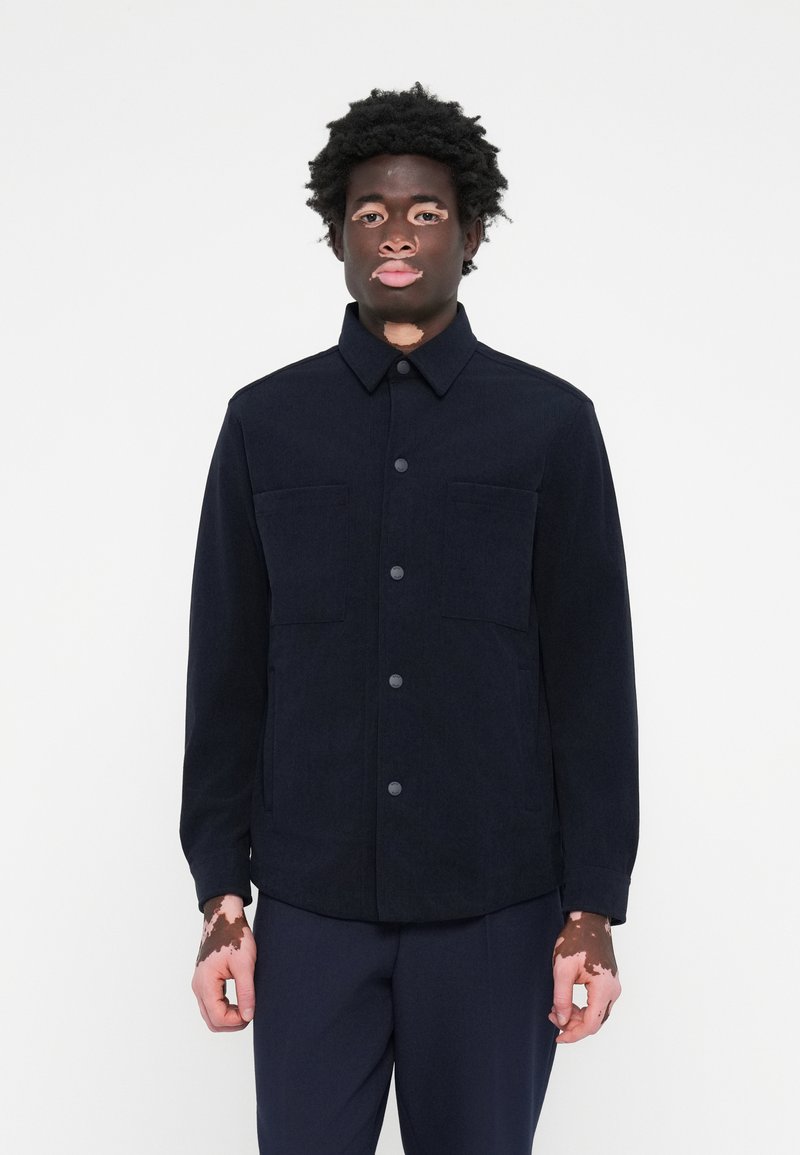 Chaqueta de trabajo azul marino en tejido estructurado. Cuenta con un cierre de botón, dos bolsillos en el pecho y un cuello. Textura suave, diseño minimalista.