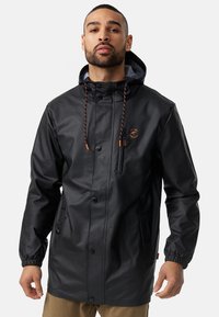 Sort vandtæt jakke med hætte, frontlommer og snoretræk. Har et orange logo og en glat, skinnende overflade.