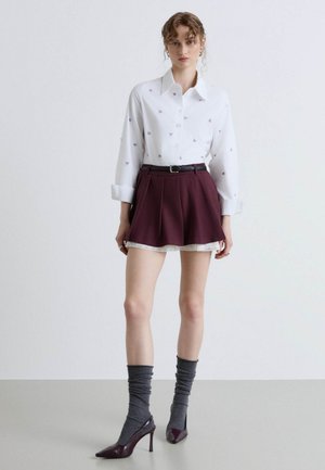 Frau trägt ein weißes Hemd mit kleinen Mustern, burgunderrote plissierte Shorts mit schwarzem Gürtel, graue Socken und burgunderrote High Heels.