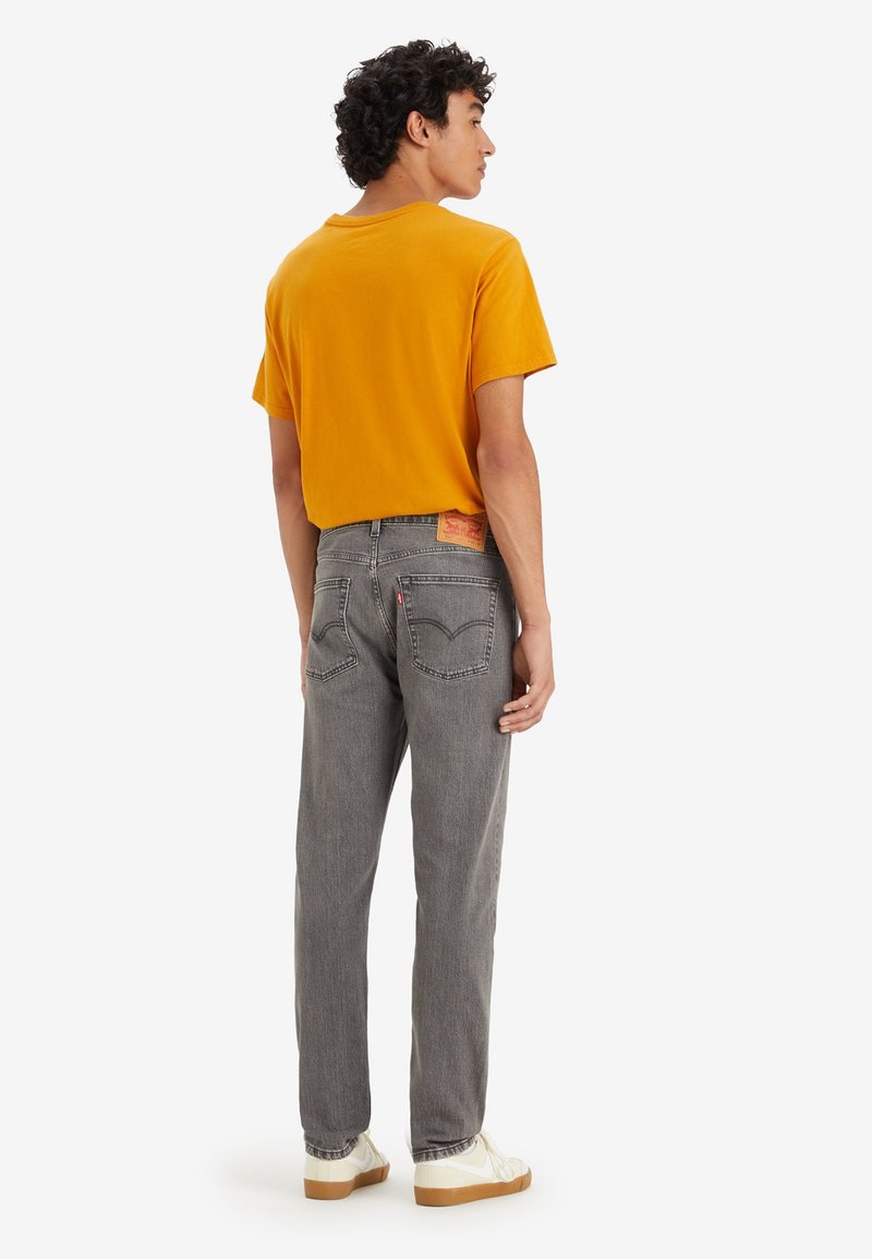 Jeans de mezclilla gris con corte recto, combinados con una camiseta de manga corta amarilla. Presenta una etiqueta de la marca en la parte posterior y calzado informal.