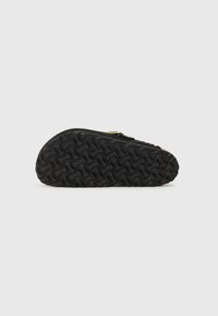 Chaussure noire avec une semelle en caoutchouc texturé, présentant un design arrondi et un dessus lisse avec une petite décoration. Détails de couture subtils visibles.