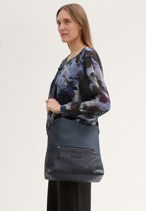 Marine leren schoudertas met een gladde textuur, voorzak met rits en subtiele logo. Bij de tas hoort een multicolore blouse.