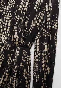 Tissu noir avec des taches beiges irrégulières, froncé et noué au centre, montrant la texture et un motif abstrait.