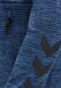 Tejido texturizado azul con un cierre negro y tres grandes flechas en forma de chevrón negras en la manga.
