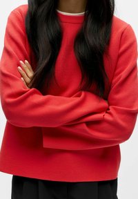 Punane kampsun ümara kaelusega ja pikkade varrukatega, mis on lõdva istuvusega. Pehme kangas nähtava tekstuuriga, kombineeritud mustade pükstega.