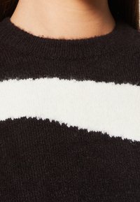 Mörkbrun tröja med ett vitt vågigt rand. Tillverkad av mjukt stickat material, med hög hals och fluffig textur. Kanten är rå och ojämn.