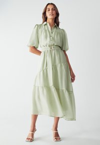 Vestido largo de color verde claro, con volantes, mangas cortas abullonadas, cierre de botones en el frente, cintura ceñida con cinturón y hebilla metálica redonda. Tejido suave y texturizado.