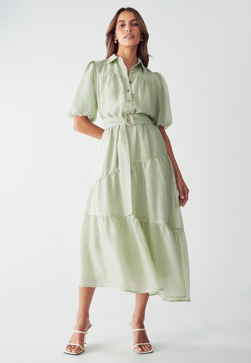 Vestido largo de color verde claro, con volantes, mangas cortas abullonadas, cierre de botones en el frente, cintura ceñida con cinturón y hebilla metálica redonda. Tejido suave y texturizado.