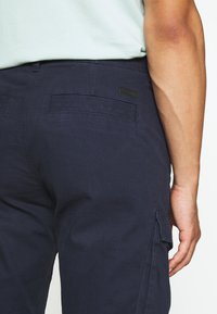 Pantalons cargo marine en mélange de coton, dotés de poches à plat, de poches latérales et d'une finition texturée. Design minimaliste avec une petite étiquette de marque.