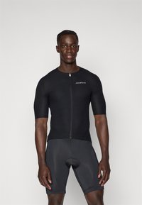 Kortermet svart sykkeltrøye med glidelås, ribbet tekstur og logo detaljer, sammen med svarte tettsittende sykkelshorts.