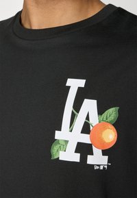 Sort bomulds t-shirt med et hvidt "LA" logo prydet med orange og grønne bladvine på venstre brystområde.